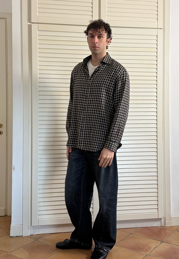 Young man standing indoors wearing a black and white checkered shirt, dark wide-leg jeans, and black shoes.