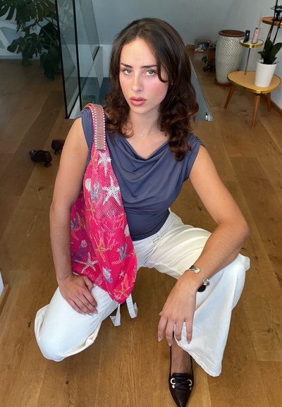 Bolso de malla rosa con patrones coloridos, llevado sobre una blusa gris, combinado con pantalones de pierna ancha blancos. Zapatos de cuero suave y pulsera de plata.