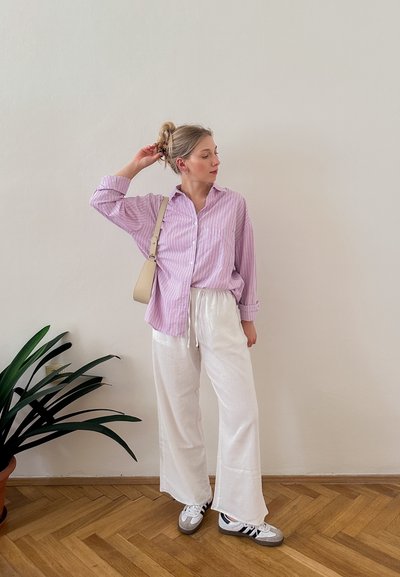 Camisa de rayas rosa claro y blanca con botones, combinada con pantalones de pierna ancha color crema y zapatillas deportivas grises. Se observa un bolso minimalista y plantas de interior.