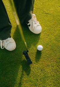 Weiße Turnschuhe mit schwarzen Streifen, graue Anzughosen und ein Putter, der über einem weißen Golfball auf grünem Gras schwebt.