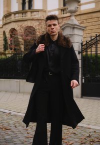 Svart kåpe med pelskrage, kombinert med svart skjorte og bukser. Utendørs omgivelser med en steinmur og en smijernsport i bakgrunnen.