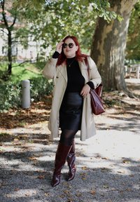 Μπεζ παλτό, μαύρο τοπ, δερμάτινη φούστα, καφέ μπότες μέχρι το γόνατο. Κρατώντας μια καφέ τσάντα και φορώντας στρογγυλά γυαλιά ηλίου. Σε εξωτερικό χώρο με φύλλωμα.