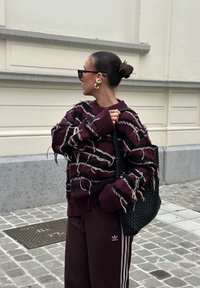 Pull bordeaux avec un motif texturé présentant des détails effilochés, associé à un sac fourre-tout tissé noir et un pantalon de sport sombre avec des rayures blanches.
