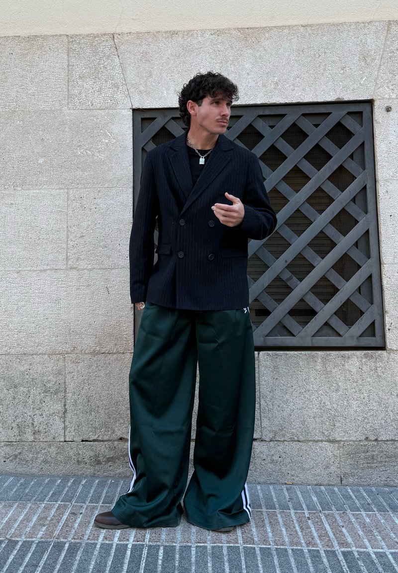 Homme debout contre un mur de pierre avec une grille en métal, portant un blazer noir croisé, un pantalon large vert foncé à rayures blanches et des baskets.