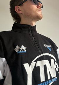Svartvit zipjacka med rutmönster och texten "YTN SPEED RACER", med blå accenter och hög krage.