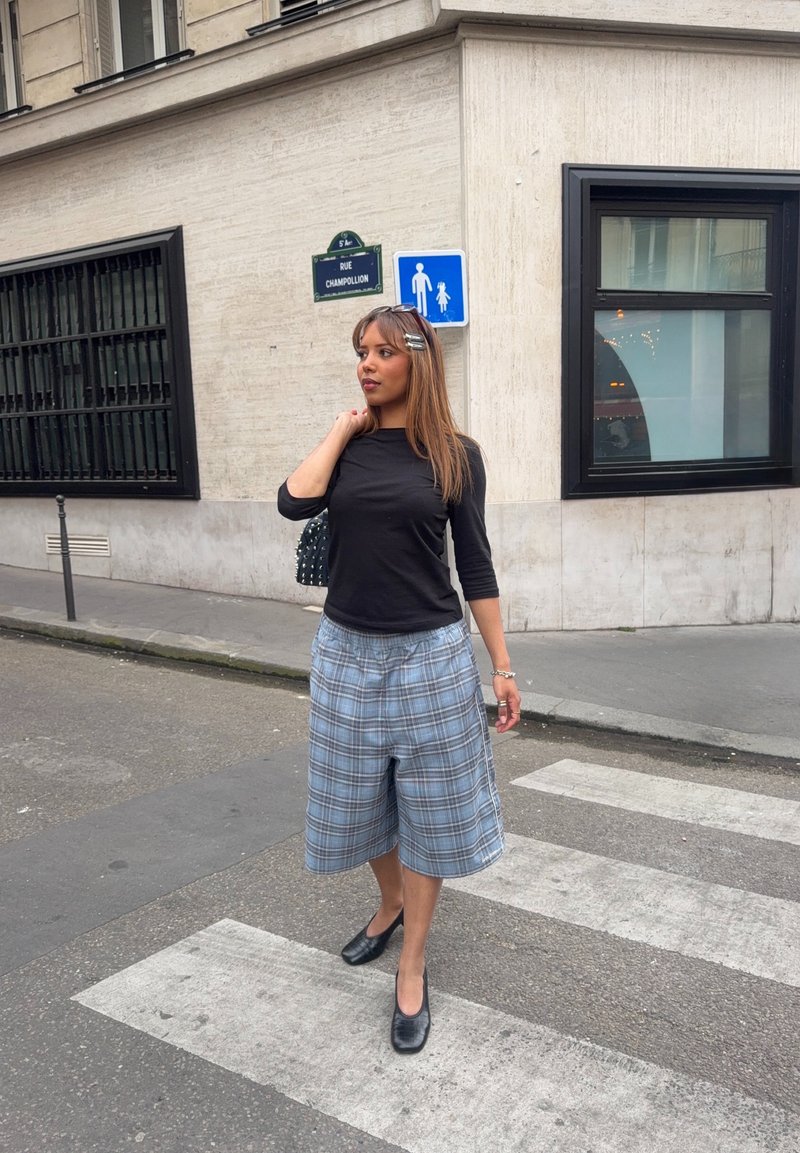 Femme debout sur un passage pour piétons, portant un haut noir, un short large à carreaux et des chaussures noires, près d'un bâtiment urbain avec des panneaux pour piétons sur le mur.