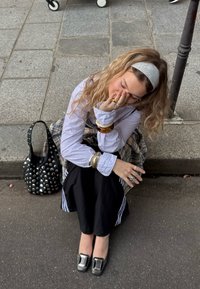 Fekete szegecses kézitáska, réteges kockás szoknya, fehér csíkos ing, ezüst és műanyag karláncok, fekete cipők ezüst csatokkal, göndör haj.