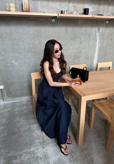 Una persona vestida con un vestido maxi azul marino está sentada en una mesa de madera clara, sosteniendo un vaso de vidrio ámbar. A su lado hay una bolsa negra.