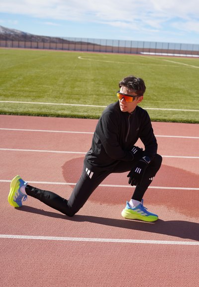Hombre con ropa deportiva negra y zapatillas de neón estirando en una pista de atletismo junto a un campo verde bajo un cielo despejado.