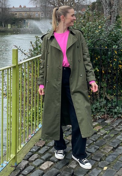 Abrigo verde sobre una camisa rosa, pantalones negros y zapatillas negras. El abrigo tiene un diseño de doble botonadura y botones.