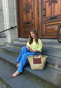 Woven straw basket bag with brown leather details, oversized yellow shirt, blue striped wide-leg trousers, and tan slide sandals on stone steps.