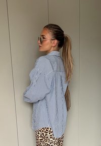 Prugasta plava i bijela prevelika košulja s leđima od joke, stilizirana s hlačama u leopard uzorku. Osoba nosi okrugle sunčane naočale i ima dugu kosu svezanu unatrag.