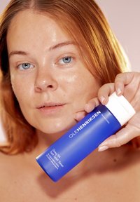 Blå cylindrisk flaska med vit pumpkork, märkt "Glow OH Dark Spot Toner," hålls av en person med ljus hy och röd hår.