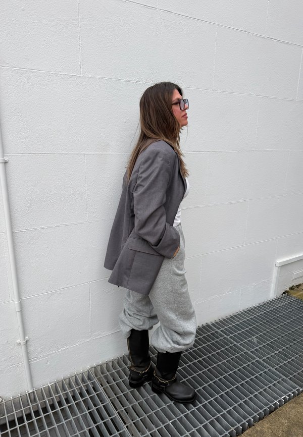 Mujer con cabello largo y castaño y gafas, de pie contra una pared blanca, vistiendo blazer gris, camiseta blanca, pantalones deportivos grises y botas negras sobre una rejilla metálica.