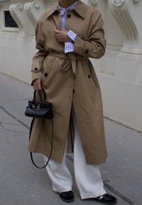 Beige trenchcoat med knapper og bælte, over en lys blåstribet skjorte. Hvide vide bukser. Lille sort håndtaske og slip-on sko.