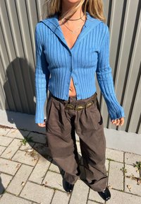 Pitkähihainen, hupullinen ribbineule vaaleansinisenä, napitettu edestä. Yhdistetty löysiin, tummanruskeisiin housuihin ja mustiin kenkiin.
