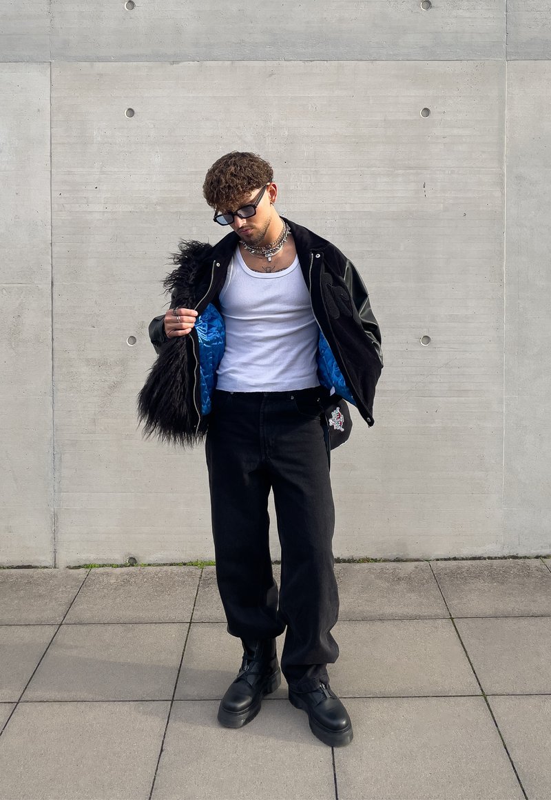 Casaco bomber preto e azul com acabamento em pele sintética, t-shirt branca, calças pretas e botas chunky pretas. Os acessórios incluem colares em camadas.