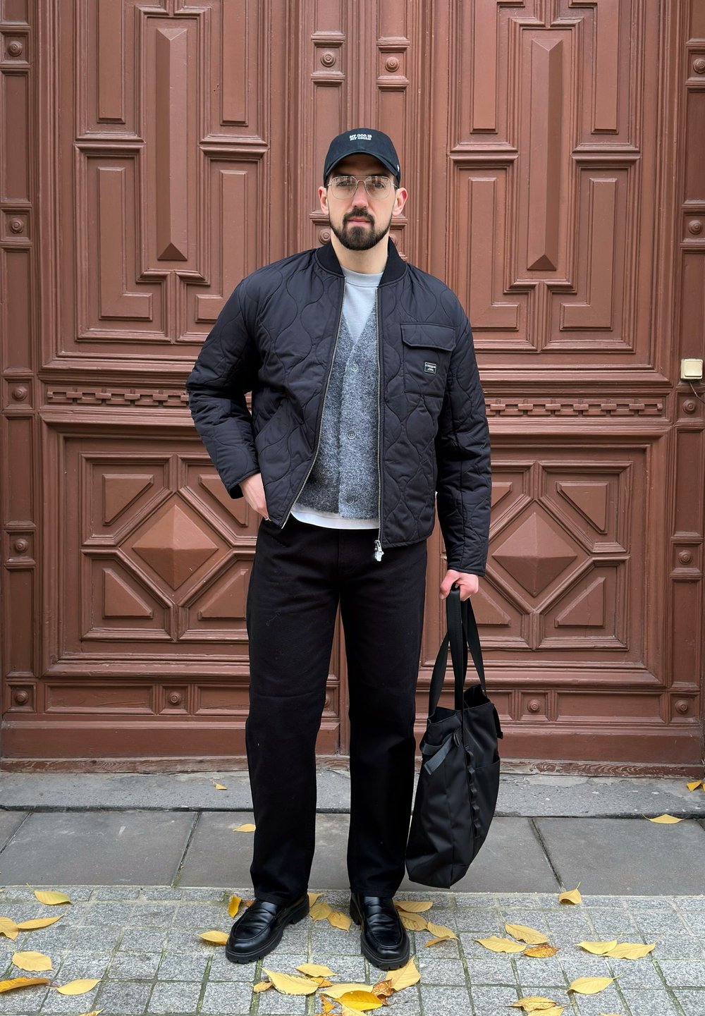 Czarna pikowana kurtka typu bomber z przednią kieszenią, szary kardigan, biała koszula, czarne spodnie i czarna torba typu tote na tle brązowych drzwi.