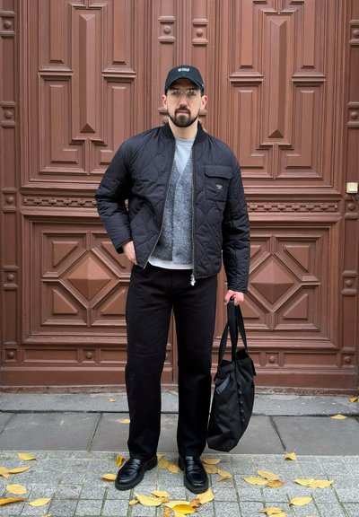 Chaqueta bomber negra acolchada con un bolsillo frontal, cárdigan gris, camisa blanca, pantalones negros y un bolso tote negro, sobre un fondo de una puerta marrón.