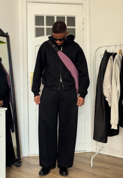 Sudadera negra con cremallera, pantalones anchos negros, zapatos negros y un bolso cruzado morado. Tejido suave y ajuste relajado con un estilo casual.