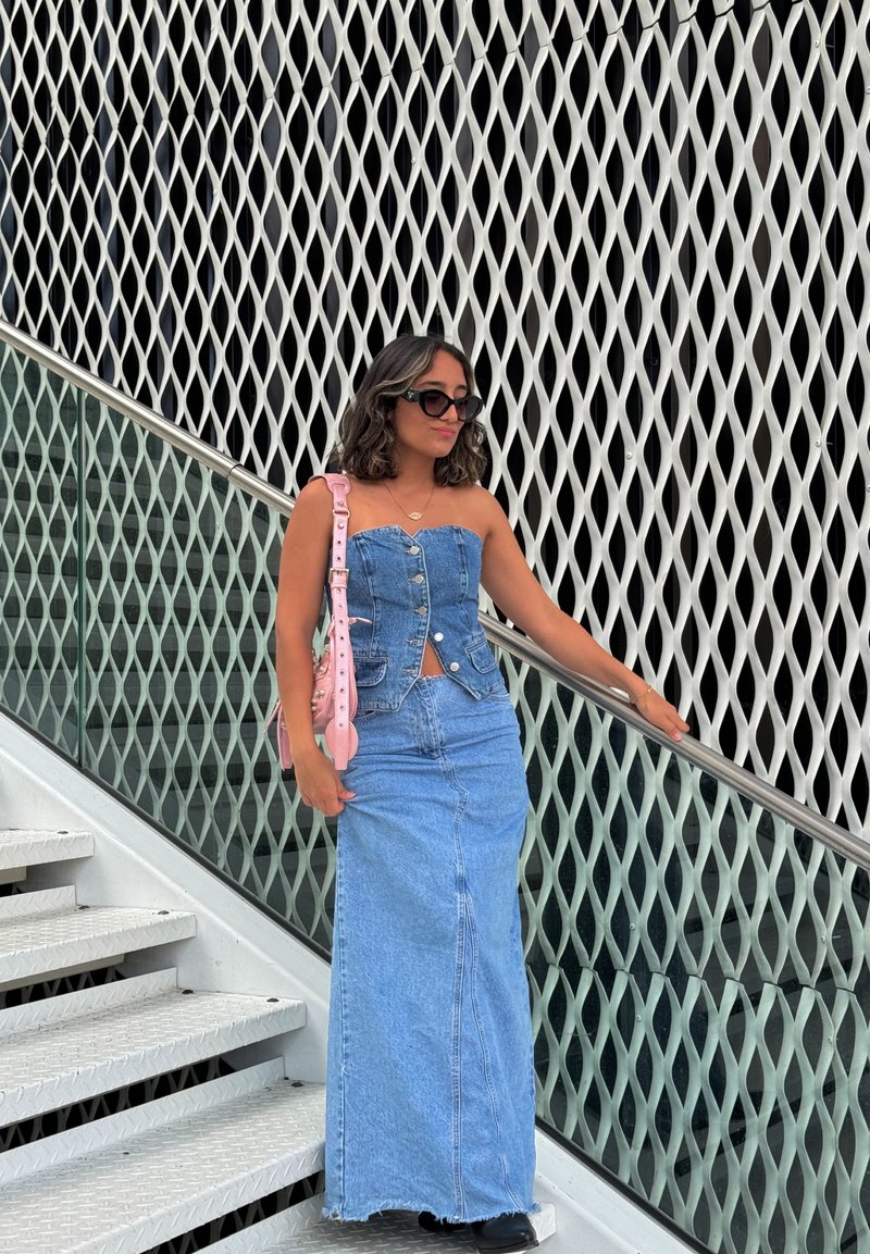 Τζιν maxi φόρεμα με εφαρμοστό κορσέ, κουμπιά και σκίσιμο; σε συνδυασμό με ροζ τσάντα ώμου και μαύρες μπότες μέχρι τον αστράγαλο. Ανοιχτό φόντο.