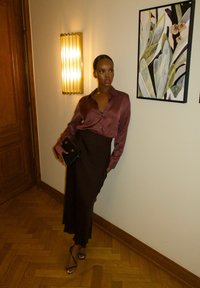 Brązowa satynowa bluzka o luźnym kroju, zestawiona z brązową spódnicą. Czarna torebka na złotym zapięciu. Ściana z teksturowanym kinkietem i ramką z roślinnym dziełem sztuki.