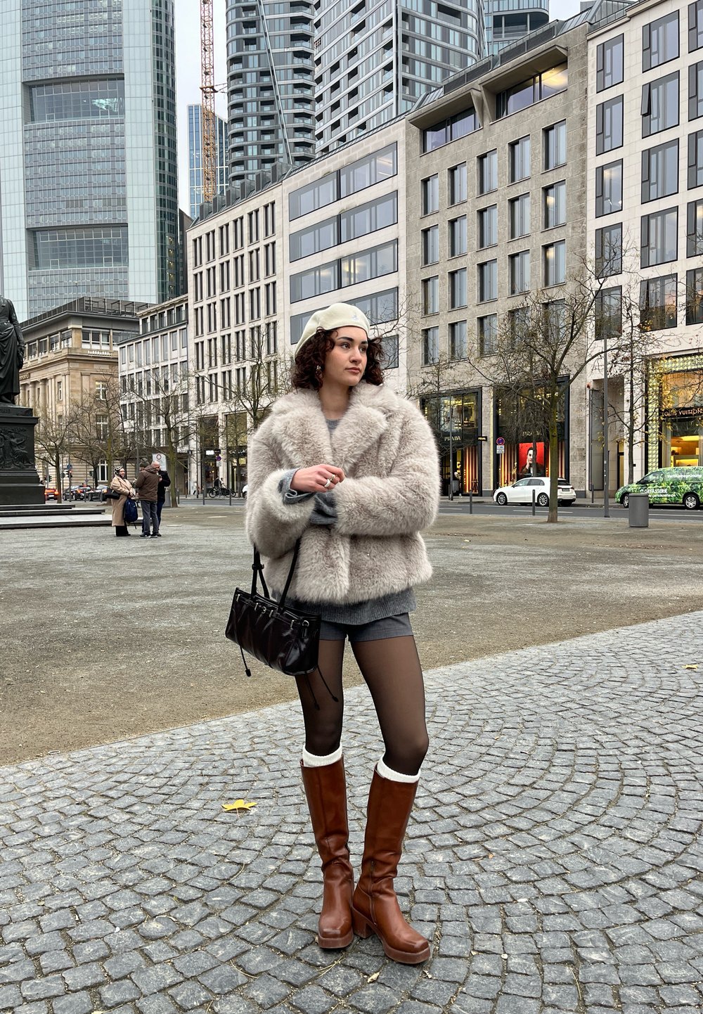 Lodden beige cropped jakke, grå top, grå shorts, brune knæhøje støvler med cremefarvede manchetter, sort håndtaske, krøllet hår; urbant miljø.