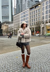 Lodden beige cropped jakke, grå top, grå shorts, brune knæhøje støvler med cremefarvede manchetter, sort håndtaske, krøllet hår; urbant miljø.