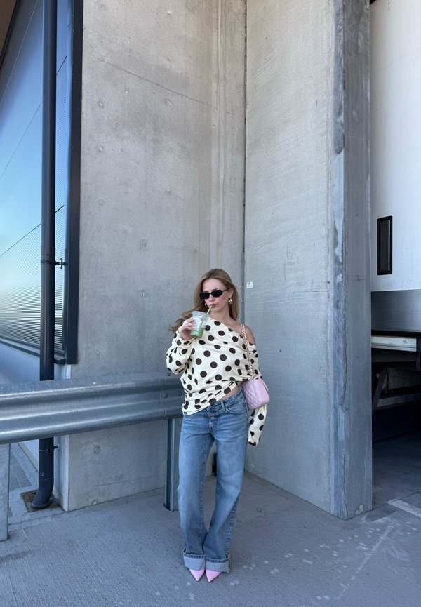 Kvinne med solbriller iført off-shoulder prikkete bluse og jeans, holder en drikke og en rosa veske, står ved en betongvegg utendørs.