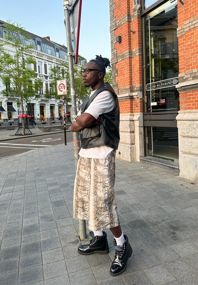 Chaleco de cuero negro sobre una camiseta blanca, falda estampada hasta la rodilla y botas negras brillantes. De pie sobre un pavimento de piedra cerca de edificios.