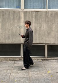 Chaqueta de cuero negra, pantalones anchos oscuros y zapatos oscuros. Persona caminando por un camino de piedra cerca de una pared de hormigón texturizado y ventanas.