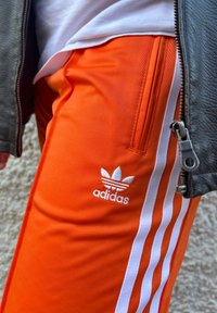 Πορτοκαλί παντελόνι Adidas με λευκές ρίγες και λογότυπο. Λείο υφή, αθλητικός σχεδιασμός, διαθέτει τσέπη με φερμουάρ και αντίθετα υφασμάτινα στοιχεία.