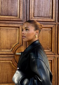 Donna con i capelli raccolti in un bun, che indossa orecchini d'oro e una giacca di pelle nera, in piedi di lato contro uno sfondo di una porta di legno ornata.