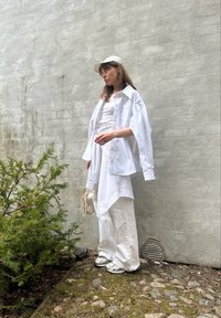 Camisa branca oversized com botões, sobre uma blusa sem mangas branca, combinada com calças largas brancas e ténis prateados. Acentuada por um boné claro.