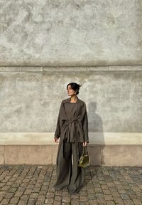 Vrouw in een losse olijfgroene outfit met een riem, die een kleine olijfgroene handtas vasthoudt, staand op een kasseienstraat tegen een gegolfde grijze muur.