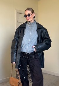 Femme portant une veste en cuir noire surdimensionnée, haut gris zippé, pantalon noir avec overlay transparent orné de fleurs pailletées, lunettes de soleil noires, tenant un sac marron.