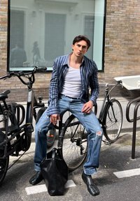 Stripete blå og hvite skjorte, hvit tanktop, slitte blå jeans, svarte skinnstøvler og en strukturert svart totebag. Syklene i bakgrunnen.