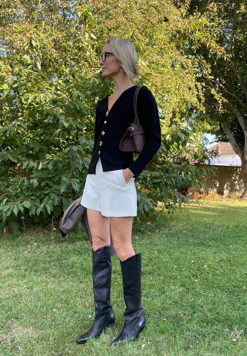 Cardigan marine con bottoni dorati, pantaloni corti bianchi e stivali alti in pelle nera. Borsa marrone con tracolla. Ambientazione all'aperto con vegetazione sullo sfondo.