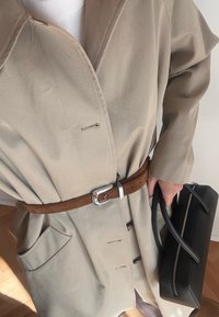 Smėlio spalvos tranšėjos paltas su rudos odos diržu. Turi šonines kišenes, V formos apykaklę ir didelius mygtukus. Juoda rankinė su užtrauktuku ir rankenomis.