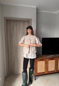 Γυναίκα με πράσινες αδιάβροχες μπότες, μαύρο παντελόνι, μπεζ φλις γιλέκο πάνω από καρό πουκάμισο και σκούφο στέκεται σε μοντέρνο δωμάτιο με ξύλινα έπιπλα.