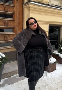 Γυναίκα που φοράει σκούρο παλτό από γούνα, μαύρο ζιβάγκο, καρό φούστα και γυαλιά ηλίου, στέκεται σε χιονισμένο δρόμο κοντά σε ξύλινη πόρτα και κίτρινο κτήριο.