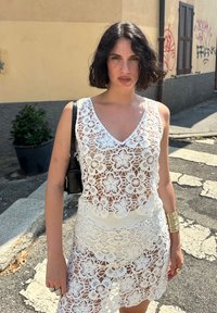 Abito bianco all'uncinetto con motivi floreali, caratterizzato da un collo a V e un design aperto. Abbinato a una borsa nera e accessori, in piedi su una strada.