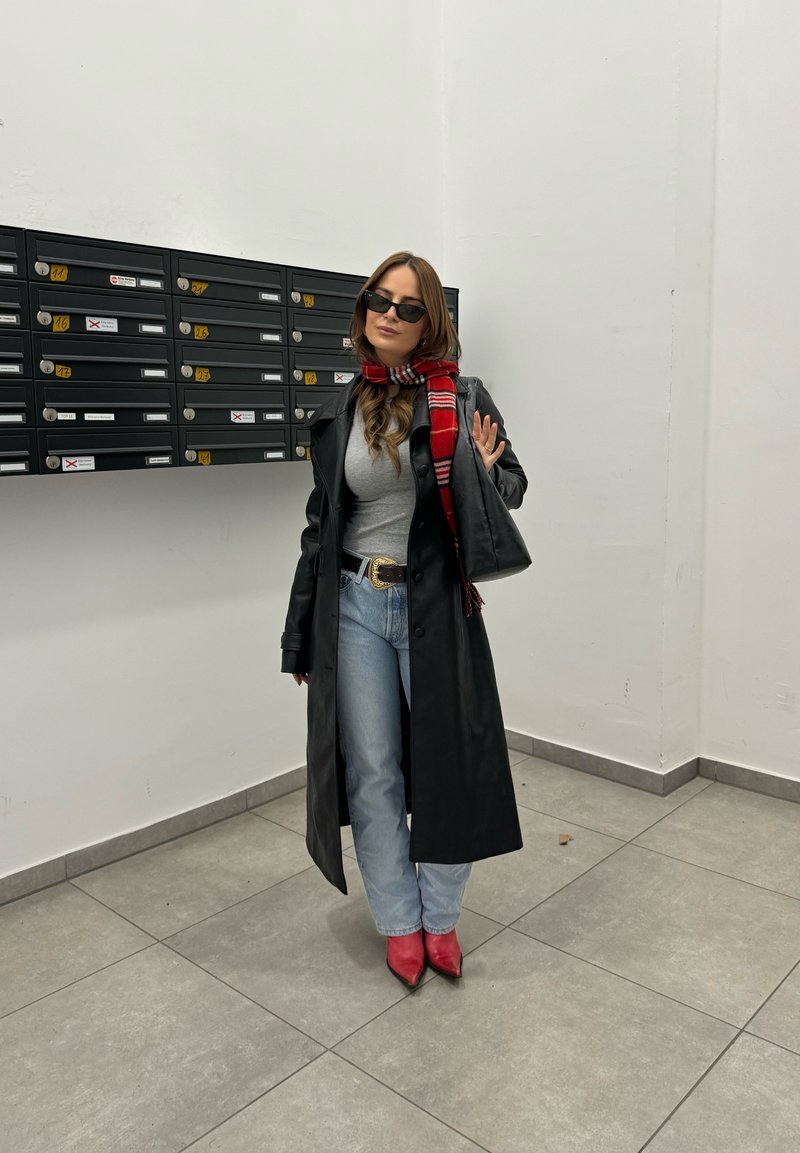 Femme portant un manteau en cuir noir, un haut gris, un jean bleu, une écharpe rouge et des bottes rouges, debout à côté d'un mur de boîtes aux lettres noires à l'intérieur.