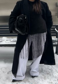 Personne portant un manteau noir et un pull, un pantalon blanc à jambes larges, des chaussures noires pointues, tenant un sac noir, debout sur un sol couvert de neige près d'un banc.