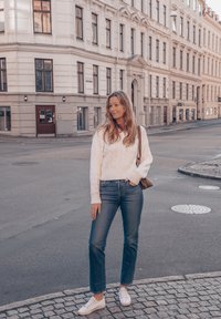 Valkoinen kaapelkudottu neule, siniset suorat farkut ja valkoiset tennarit. Seisoo kivikatu kadulla ruskean olkalaukun kanssa.