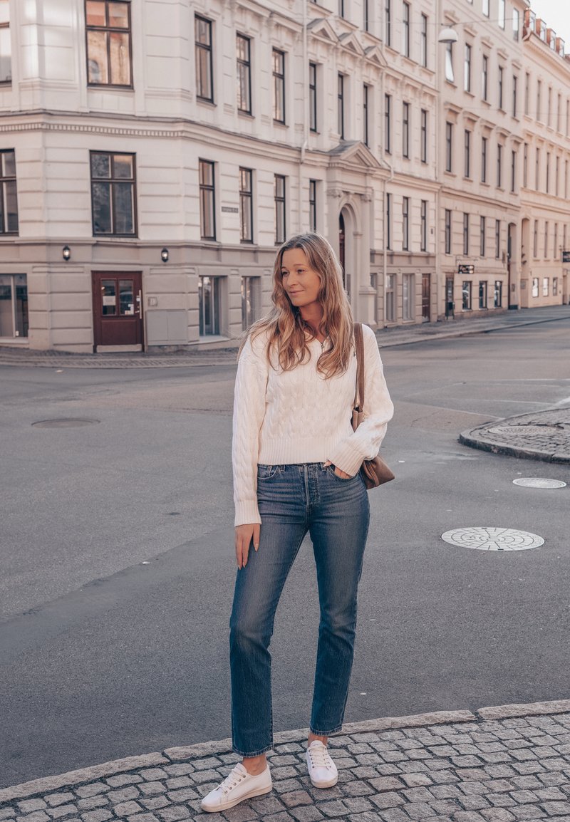 Bijela pletena veste, plave ravne traperice i bijele tenisice. Stoji na kaldrmi s smešnom torbom preko ramena.