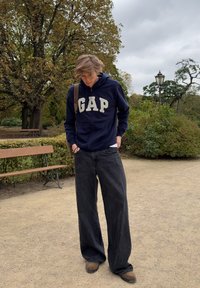 Marine zip-up hoodie med "GAP" i cremefarvede bogstaver, parret med brede sorte denimbukser. Omgivelsen har bænke og grønne områder i en park.