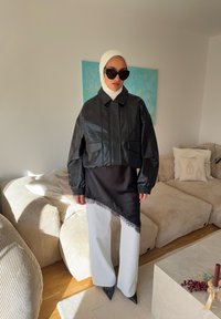 Veste courte en cuir noir avec deux poches frontales, superposée sur un haut asymétrique noir avec bordure en dentelle, assortie à un pantalon blanc à jambes larges.