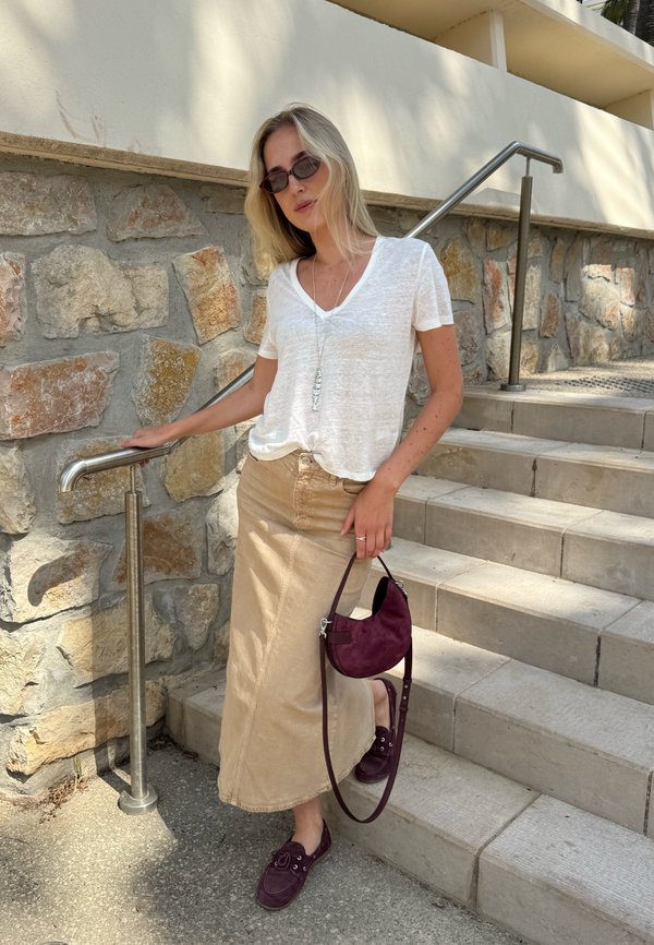 Žena s blond vlasmi, oblečená v biele tričko, béžovú dlhú sukňu, tmavé slnečné okuliare, drží fialovú tašku, stojí na schodoch pri kamennom múre s kovovým zábradlím.