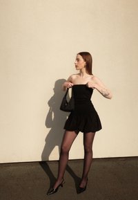 Haut noir sans bretelles associé à une jupe noire évasée. La silhouette est mise en valeur par des collants semi-opaques noirs et des talons noirs pointus.